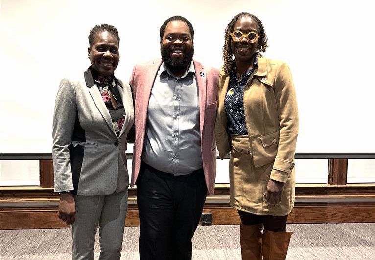 3 people standing together and smiling in a large room. John Anthony Fuller is in the center wearing a pink blazer, with a woman in a gray suit on his left and a woman in a tan jacket and boots on his right